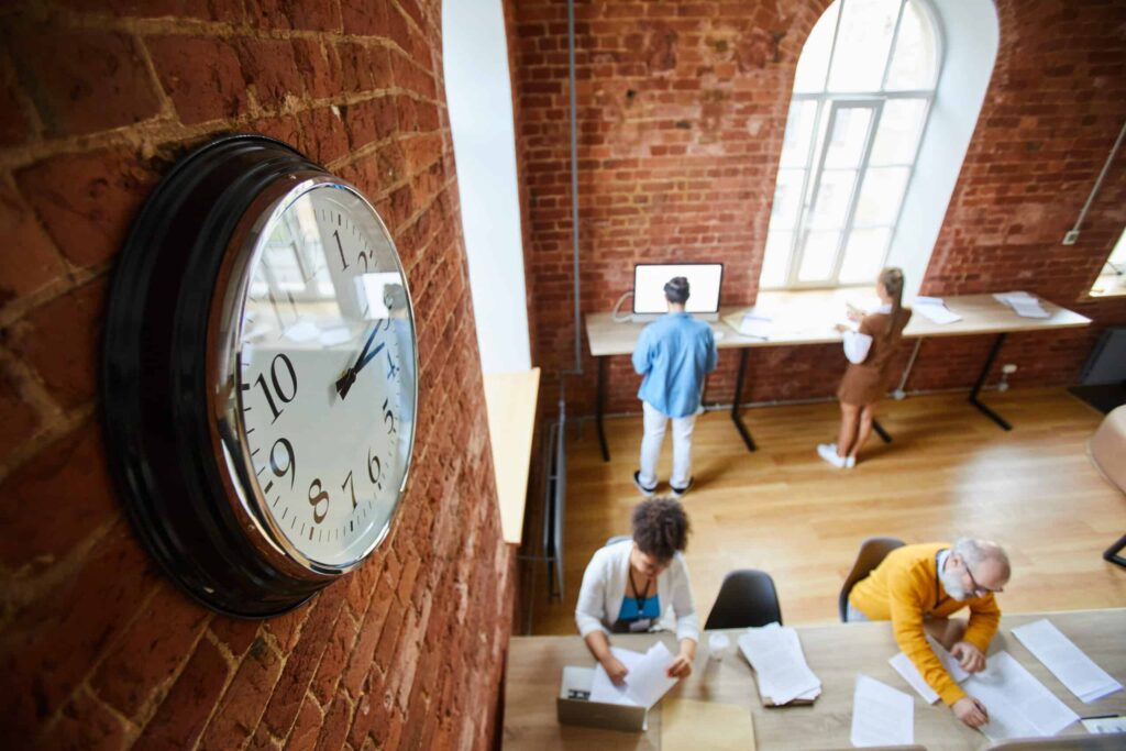 Modern office workspace with colleagues collaborating and a clock on the brick wall.