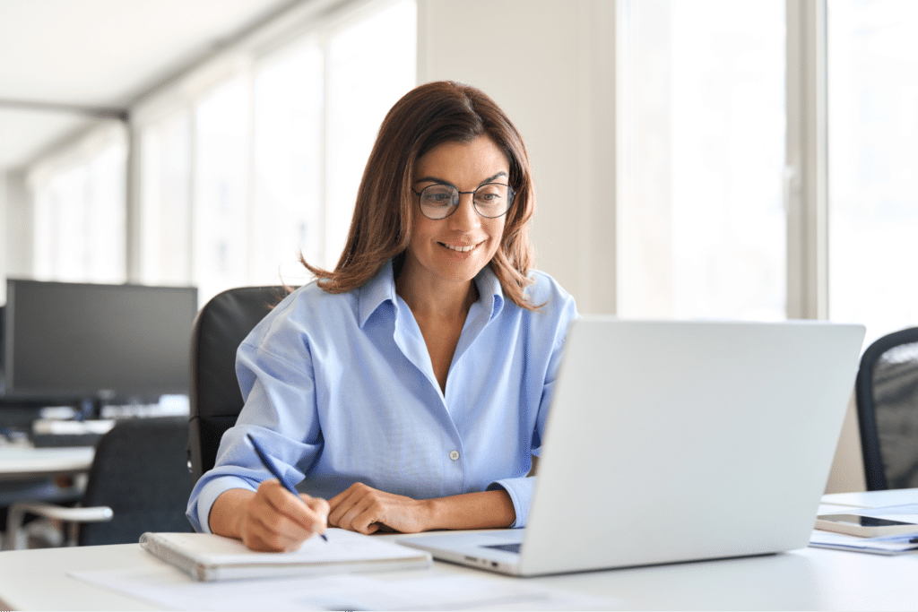 Professional woman focused on laptop, taking notes in a bright office setting.