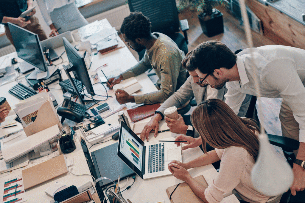 Collaborative office environment with diverse team working on laptops and discussing projects.